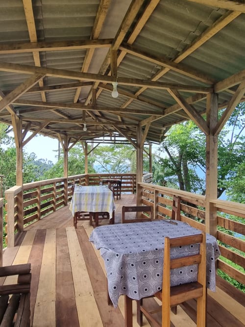 Deck de madeira com vista para o mar em Vila Paraíso, São Tomé. Mesa com toalha estampada e cadeiras sob telhado de metal ondulado