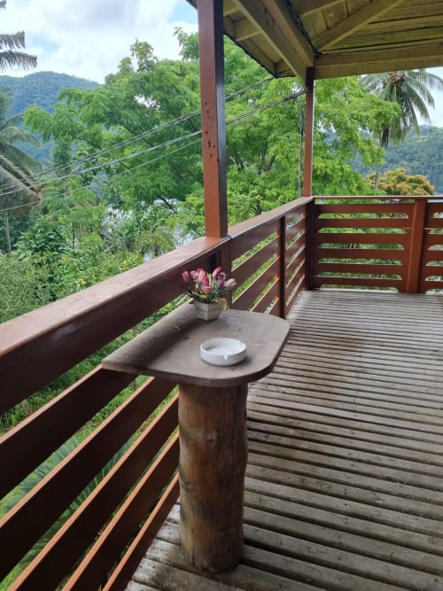 Varanda com deck de madeira, flores rosa, cinzeiro branco e vistas exuberantes de montanhas tropicais em Vila Paraíso, São Tomé
