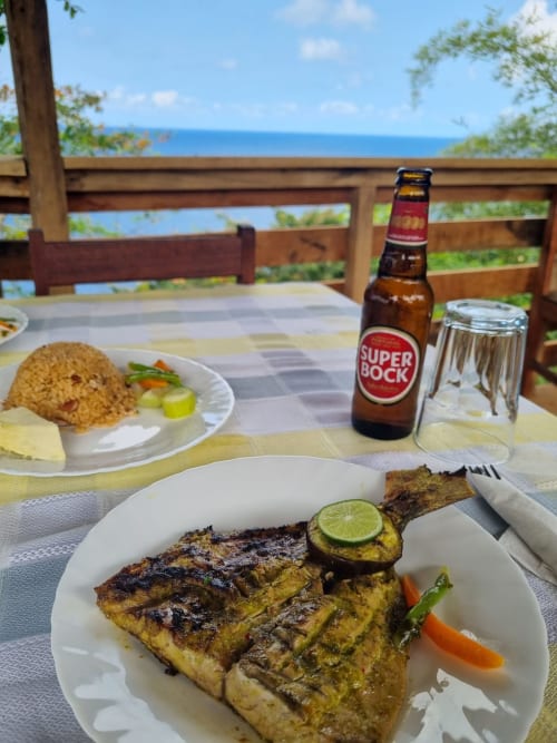 Peixe grelhado com limão e vegetais, cerveja e arroz em restaurante à beira-mar em São Tomé com vista para o oceano