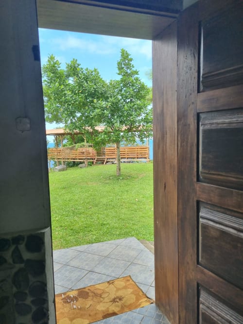 Porta de madeira aberta revelando varanda com vista para o mar na Equador Houses, quarto Vila Paraíso em São Tomé com jardim exuberante e deck de madeira