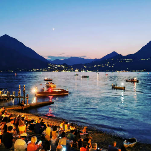Twilight gathering on Lake Como beach with illuminated boats and mountains, Villa Vista Lago di Como Bellano