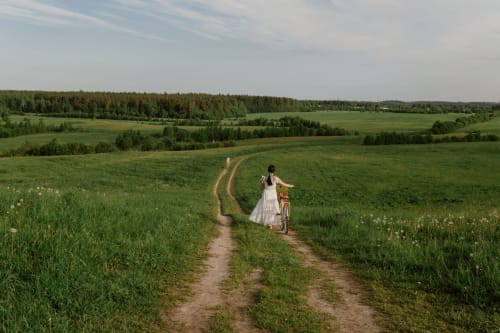 Woman in white dress pushing bicycle on rural dirt path through green fields near Chateau Haute Roche, Oudon, France