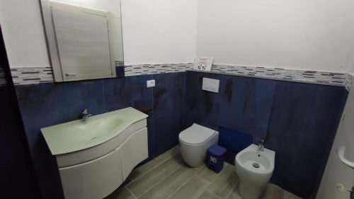 Bagno moderno nella camera Yaki Domo Platinum a Cagliari, con lavabo bianco, wc, bidet, piastrelle blu scuro e finiture bianche