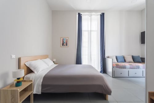 Camera moderna e luminosa a Darna Rooms Napoli con letto in legno, biancheria grigia, tende blu e seduta integrata alla finestra