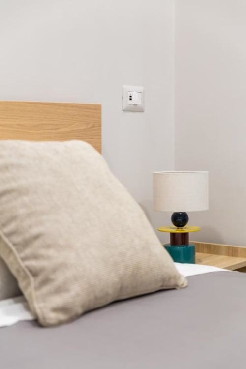 Dettaglio camera Darna Rooms Napoli: letto in legno, cuscino bianco, lampada moderna colorata, presa elettrica
