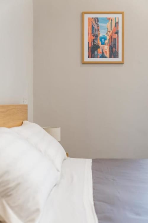Camera da letto minimalista presso Darna Rooms Napoli con opera d'arte incorniciata che ritrae una vibrante scena di strada italiana sopra il letto in legno