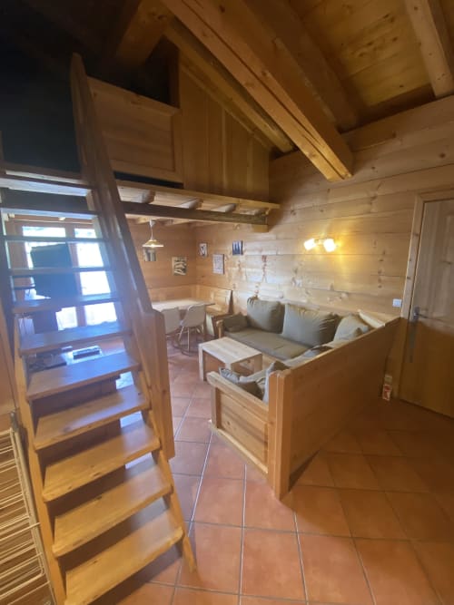 Intérieur de chalet en bois avec échelle de mezzanine, séjour avec canapé, coin cuisine et plafond à poutres apparentes à La Ferme des