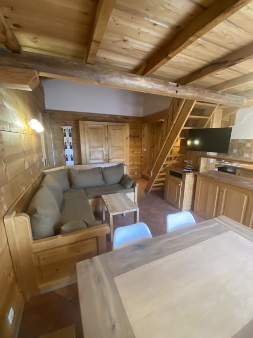 Intérieur d'appartement en bois à La Ferme des Georgières avec table à manger, canapé gris, cuisine, TV et plafond avec poutres apparentes