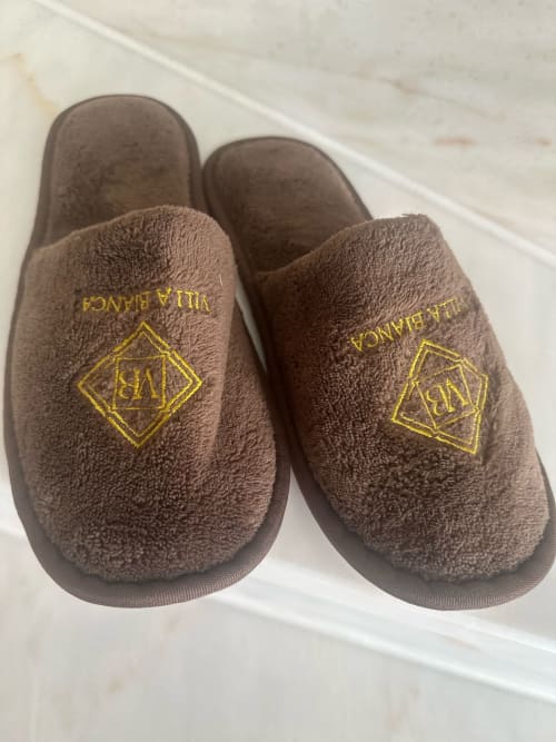 Villa Bianca-pantoffels in taupe met goudgeborduurde logo op marmeren oppervlak