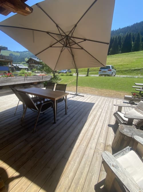 Terrasse ensoleillée avec grand parasol et mobilier en bois dominant une prairie alpine verdoyante et des montagnes boisées à La Ferme des