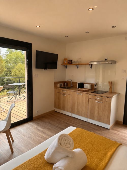Kitchenette avec meubles en bois, TV, étagères et lit à couverture jaune au Gîte l'Harmonie du Causse