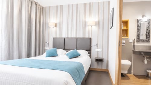 Chambre moderne de l'Hôtel d'Orléans avec tête de lit grise, accents bleus, papier peint rayé et salle de bain attenante