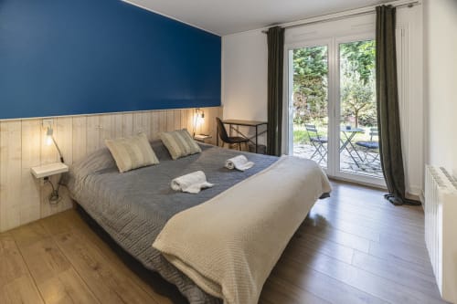 Chambre double lumineuse à l'Hôtel De La Bonnheur avec mur d'accent bleu, tête de lit en bois, balcon avec vue sur le jardin et décor moderne