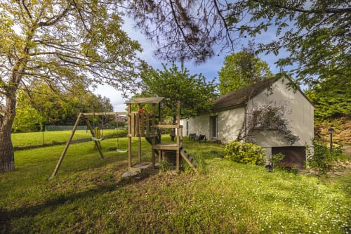 Vue du jardin de l'appartement Hôtel De La Bonnheur avec aire de jeux en bois, chalet blanc, pelouse verdoyante et arbres environnants