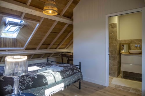 Chambre mansardée à la Cour Du Liège avec lucarne, poutres apparentes, lit en métal et salle de bains attenante aux carrelages en pierre