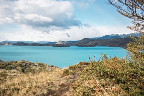 Lac turquoise avec montagnes enneigées, végétation clairsemée et ciel nuageux