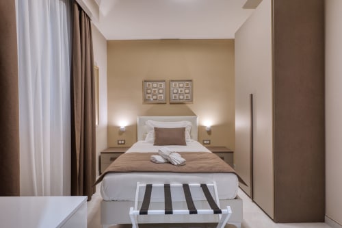 Camera da letto moderna a Marea Sliema con parete in rilievo beige, biancheria bianca, plaid marrone, applique da parete e panca a righe