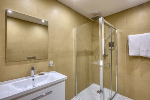 Bagno moderno a Marea Sliema con piastrelle beige, lavabo bianco, specchio e box doccia in vetro