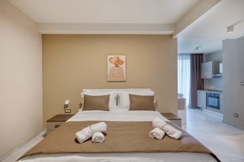 Camera da letto elegante Marea Sliema con pareti beige, biancheria bianca, cuscini marrone, asciugamani arrotolati e angolo cottura visibile