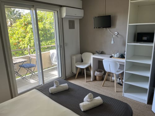 Modern hotel room at Hôtel l'Orangeraie Lavandou with desk, balcony garden view, AC unit, flat-screen TV, and white shelving.