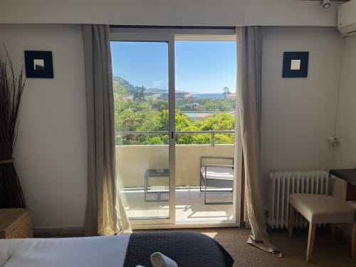 Bright hotel room with glass doors opening to balcony overlooking gardens and sea at Hôtel l'Orangeraie Lavandou