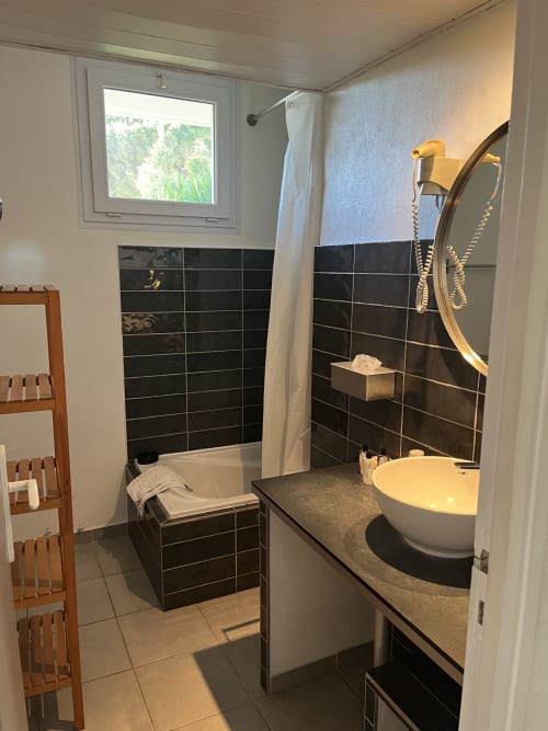 Modern bathroom at Hôtel l'Orangeraie Lavandou with black tile walls, round white sink, bathtub, and garden view window