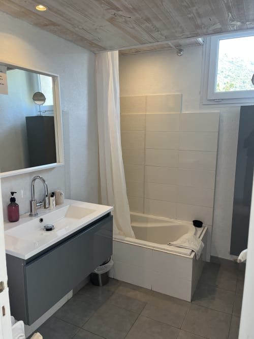 Modern bathroom at Hôtel l'Orangeraie Lavandou with dark vanity, white basin, bathtub with shower curtain, and tiled flooring
