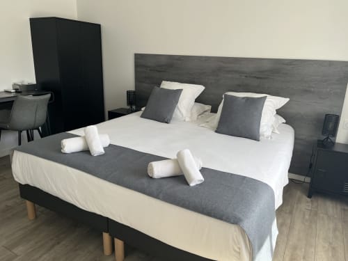 Modern hotel bedroom at Hôtel l'Orangeraie Lavandou with queen bed, gray headboard, white bedding, and rolled towels