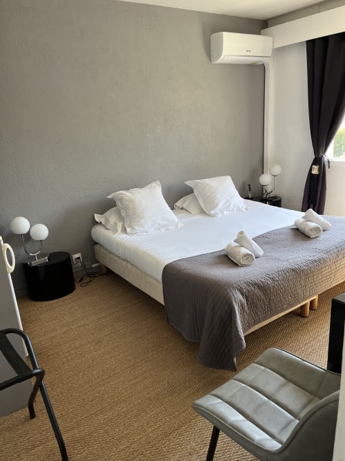 Modern hotel room at Hôtel l'Orangeraie Lavandou with white pillows, gray bedding, rolled towels, wall-mounted AC, and dark