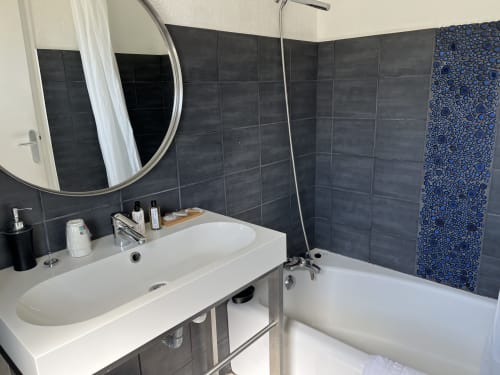 Modern bathroom at Hôtel l'Orangeraie Lavandou with dark tiled walls, round mirror, white sink, bathtub with blue towel, and