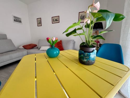 Tavolo giallo brillante con vaso turchese e pianta in vaso nella zona living della Costabella Guest House, divano grigio visibile