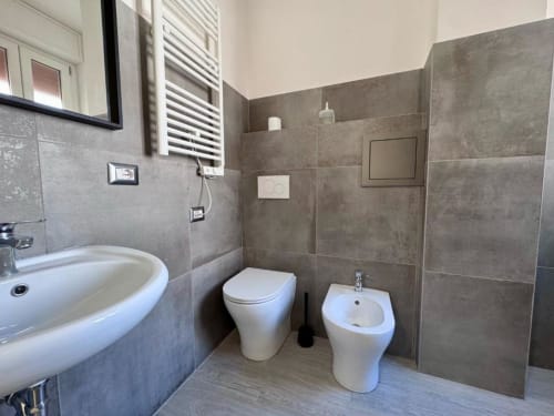Bagno moderno della Costabella Guest House con pareti in piastrelle grigie, wc bianco, bidet e lavabo a colonna con specchio