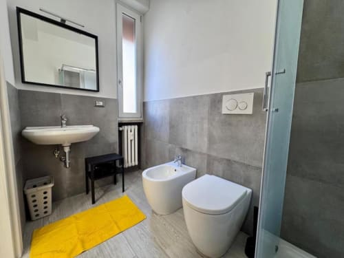 Moderno bagno della Costabella Guest House con bidet, wc, lavabo su colonna, specchio e tappetino da bagno giallo su pavimentazione in piastrelle