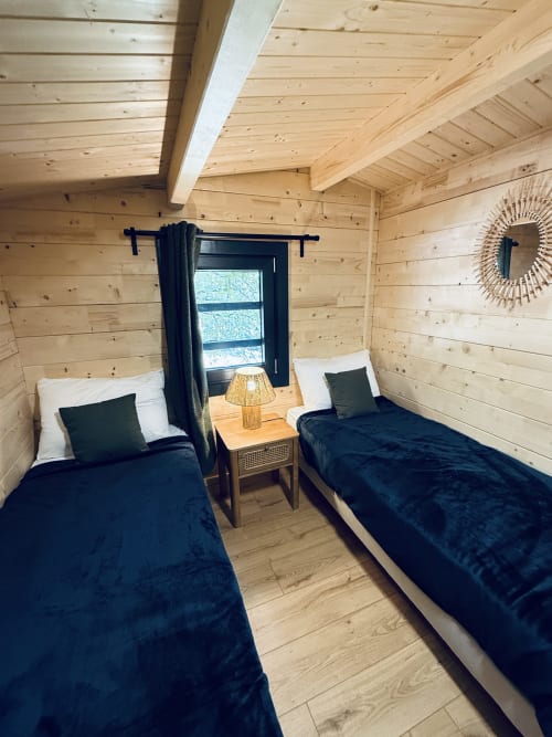 Cozy twin bedroom in wooden lodge with dark blue bedding, nightstand lamp, and window view at Cabanes du Belvédère,