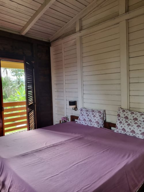 Quarto na Equador Houses em São Tomé com lençóis roxos, almofadas florais, venezianas de madeira e vista para o jardim tropical