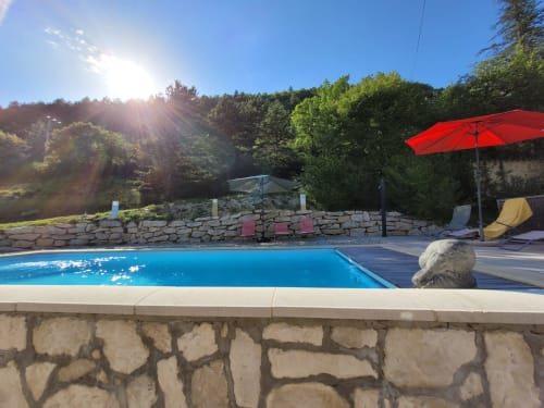 Après-midi ensoleillé à la piscine du Domaine de la Méouge avec parasol rouge, chaises longues, terrasse en pierre et montagnes boisées
