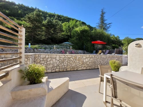 Terrasse ensoleillée du gîte Domaine de la Méouge avec murs en pierre, transats et vue sur les montagnes boisées