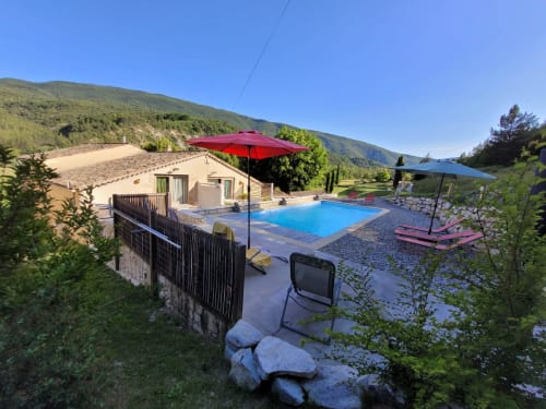 Vue montagne du gîte Domaine de la Méouge avec piscine bleue, parasol rouge, transats et collines verdoyantes