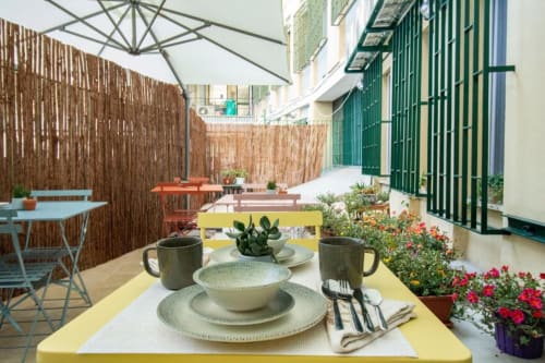 Tavolo giallo brillante con coperto nel cortile del B&B Epimoni a Catania, circondato da vasi di fiori e staccionata in legno