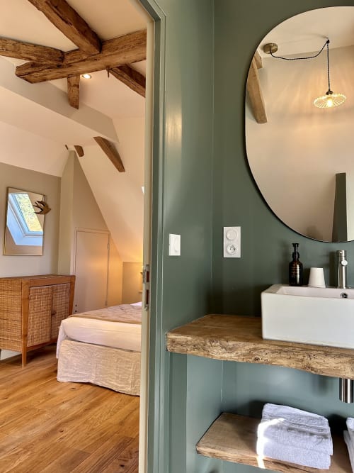 Salle de bains attenante moderne aux murs verts, étagères rustiques en bois, miroir rond et vasque au Manoir du Rigouneix