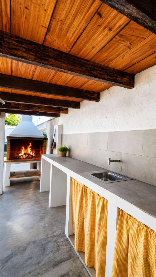 Cocina exterior de La Caseta de Ròtova con chimenea, vigas de madera, encimeras blancas y cortinas amarillo dorado