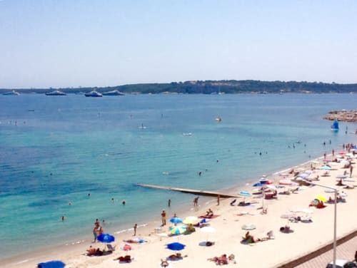 Cannes Palmenstrand mit kristallklarem türkisfarbenem Wasser, Sandstrand, Sonnenanbeter und ankernden Booten an der Französischen Riviera