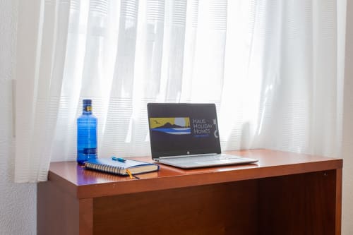 Área de trabajo en Haus Holiday Homes, Cartagena, con laptop, cuaderno y botella azul junto a ventana con cortinas translúcidas