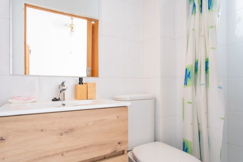 Baño moderno con mueble de madera, azulejos blancos y cortina de ducha con motivos de hojas verdes en Villa Hammely, Cartagena