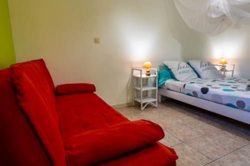 Chambre lumineuse aux Gîtes Bagatelle à Sainte-Rose avec fauteuil rouge, lit blanc aux accents turquoise et éclairage chaleureux