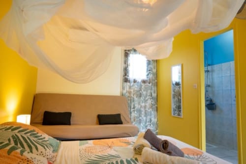 Chambre colorée aux Gîtes Bagatelle : murs jaunes, voile de lit aérien, canapé beige et salle d'eau carrelée bleue à Sainte-Rose