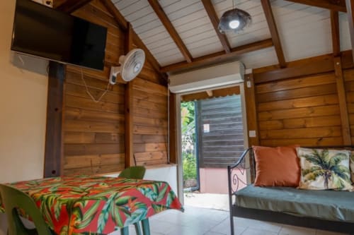 Cozy wooden cottage interior at Gîtes Bagatelle, Guadeloupe with bed, sofa, TV, fan and terrace access