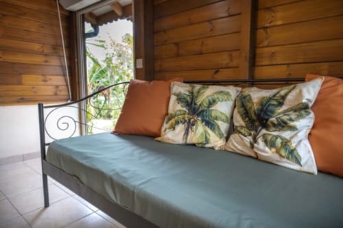 Cozy cottage bedroom at Gîtes Bagatelle in Guadeloupe with wooden walls, metal bed, tropical cushions, and garden view