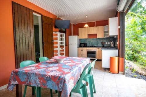 Open-plan kitchen and dining area with orange walls, green chairs, and colorful tablecloth at Gîtes Bagatelle in Guadeloupe