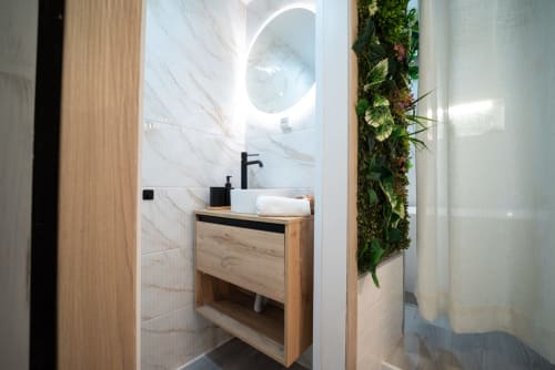 Baño moderno en apartamento Luxer Holidays con tocador de madera, azulejos de mármol, espejo circular y pared de plantas vivas